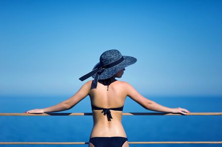 a girl in a black hat and a swimsuit stands at the railing on the roof of the hotel and looks awayの写真素材
