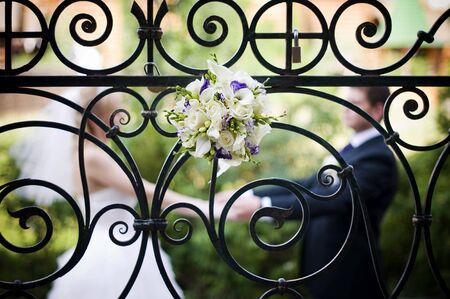 A brides bouquet is inserted into an elegant wrought-iron fence, behind which the newlyweds hold hands in defocus. grainの写真素材