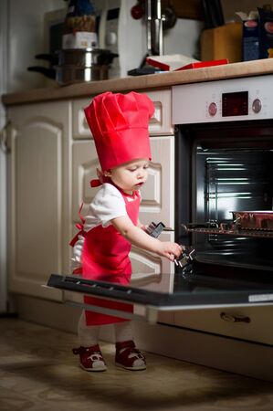 baby girl in a red cook suit plays with kitchen utensils near the ovenの写真素材