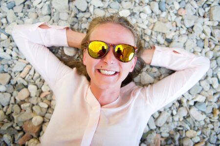 a young woman in a white shirt and glasses lies on a pebble and smiles broadly, and a photographer is reflected in her sunglassesの写真素材