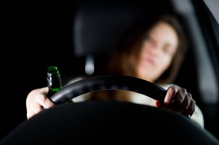 The girl leaned back in the driver's seat and holds her hands on the steering wheel, in one of her hands she has a bottle of alcoholの写真素材