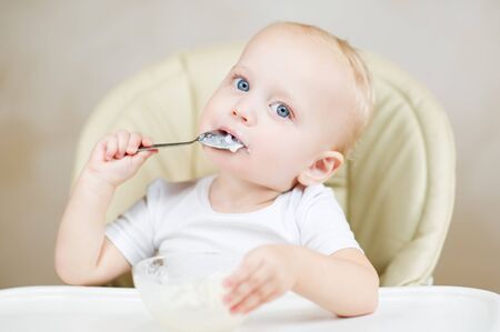 A little girl eats porridge with a spoon, sitting on a feeding chair with a white table and is thinking about something. View from aboveの写真素材