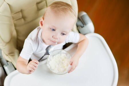 A little girl eats porridge with a spoon, sitting on a feeding chair with a white table and is thinking about something. View from aboveの写真素材