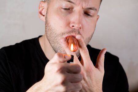 unshaven men holds a sausage in his mouth in the manner of a cigar and is about to light it