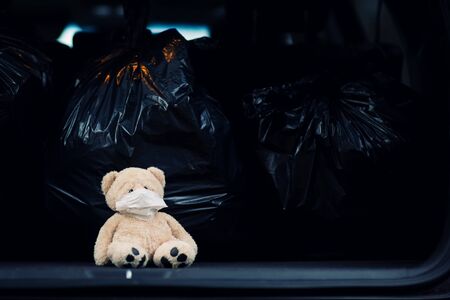 a small teddy bear in a medical mask sits in the trunk of a car with garbage bagsの写真素材