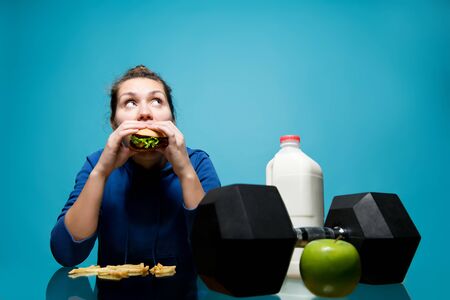 young woman eating a burger, preferring it to a dumbbell and milk with an apple, blue background