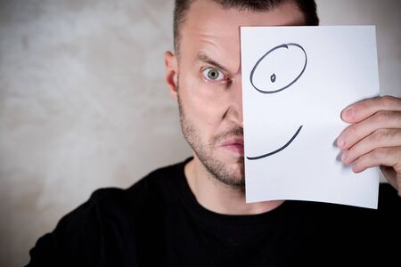 angry man covers half his face with a piece of paper with a painted smile. copy spaceの写真素材