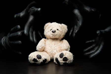 many black-gloved hands reach for a teddy bear from the dark, close up