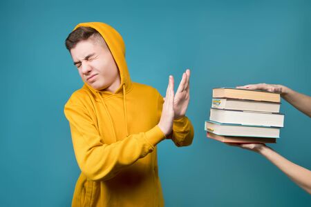 A girl holds out a stack of books to a young guy in a yellow sweatshirt on a blue background, and the guy tries to push it away from him with his handsの写真素材