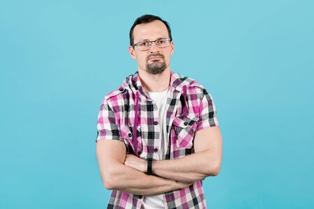 A young man with a beard in glasses and a clerical shirt folded his arms over his chest on a blue...