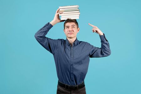 A teenager in a blue shirt with discontent holds a stack of books on his head and points a finger at it.の写真素材