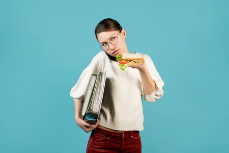 A girl with glasses with documents under her armpit is talking on the phone and is about to eat a sandwich. Quick snack concept, food on the go.の写真素材