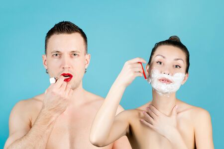 a man paints his lips with red lipstick and a girl shaves her face with a razor with shaving foam on a blue background. Theme of gender, roles in societyの写真素材