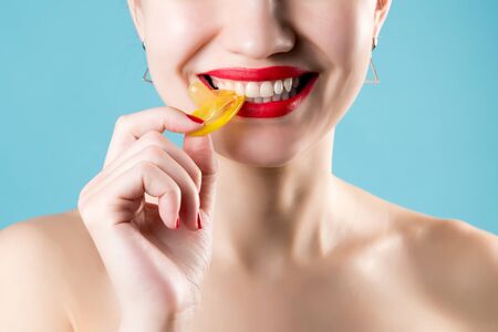A girl with red lipstick and red nails on a blue background smiles, bites and pulls a yellow condom. Close-upの写真素材