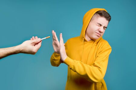 a teenager in a yellow sweatshirt on a blue background shows disgust when they hold out a cigarette,...