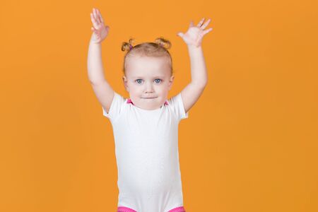 Little girl with ponytails pulls her hands up. Isolated on orange yellow background.の写真素材