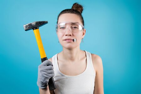 Young woman squints, holding a hammer and nails in her mouth. Isolated on blueの写真素材