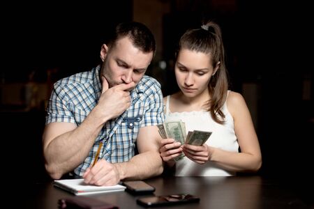 The guy and the girl are counting the money and looking thoughtfully at the numbers in the notebook. Financial difficulties in a young familyの写真素材