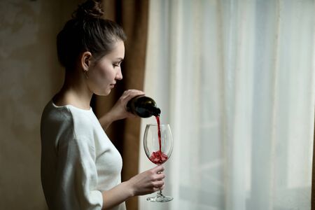 Young woman pours red wine into a large glass near a curtained window in the hallwayの写真素材