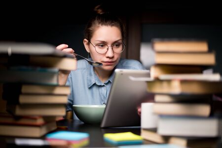 A student girl eats and rests from study - she watches the series on the tablet, and around there is a large amount of training materialsの写真素材
