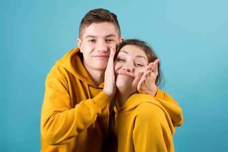 brother squeezes the cheeks of an older sister, fooling around on a blue background. Close-up, teenagers in yellow hoodiesの写真素材