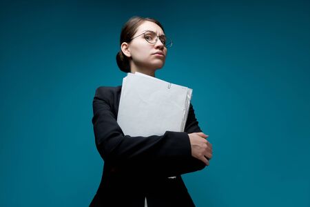 A young woman with glasses looks down at the camera, holding documents. Female boss on a blue backgroundの写真素材