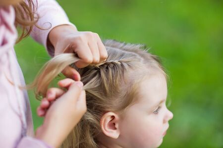 Weaving braids for a child close-up. Hands of a woman who weaves a braid from the hair of a blonde girl, close upの写真素材
