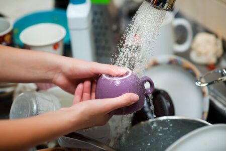 washing cups under running water against the background of a sink full of utensilsの写真素材