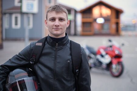 Smiling biker holds a helmet on the background of a motorcycle in the evening in the parking lotの写真素材