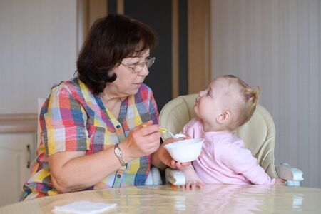 General view of the grandmother and granddaughter in the process of feeding yogurt. Nanny and little child eat food at the table in the kitchenの写真素材