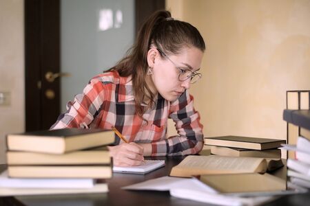 A girl prepares for exams by reading and taking notes. She is sitting at a table with many books....