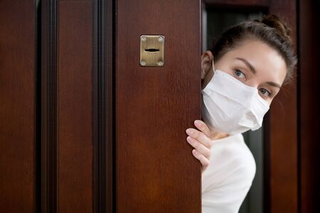 A woman in a medical mask looks at the street through an ajar front door. Inability to go outside during quarantineの写真素材