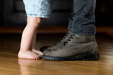 A little girl with bare feet is standing in front of her father in the hallway. dad is shod and is about to leave somewhere. Close up, no face, only legsの写真素材