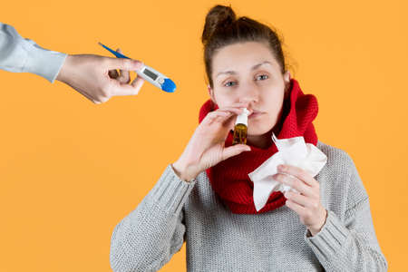 a sick girl is offered a thermometer to measure temperature. Sick girl wrapped in a scarf and sprinkles medicine in her nose. Isolated on yellow backgroundの写真素材