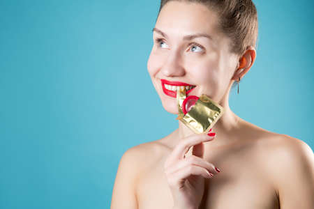 Girl playfully holds the edge of the wrapper of a red condom with her teeth, smiling. The girl has red lipstick and nails. Blue background, girl without clothesの写真素材