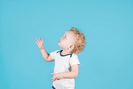Two-year-old curly-haired baby girl reaches for something, turning sideways to the camera. Isolated on blueの写真素材