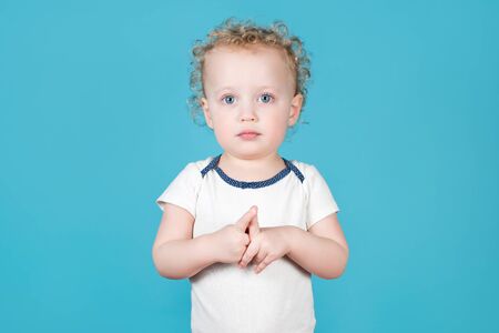 Two-year-old curly baby girl carefully looks at the camera, hands clasped together. Isolated on blueの写真素材