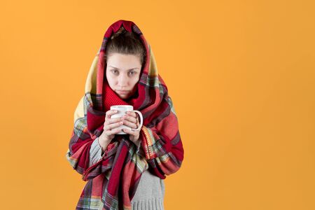 A sickly-looking girl wrapped herself in a warm plaid and looks tiredly forward with a mug of tea in her hands. Isolated on an orange-yellow background.の写真素材