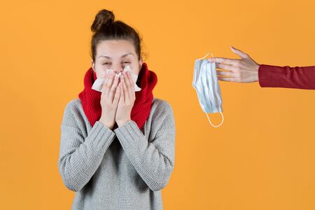 A girl with symptoms of the virus and a cold is offered to wear a mask. A hand holds out a medical mask to a girl who is actively sneezing and coughing. Isolated on orange background.の写真素材