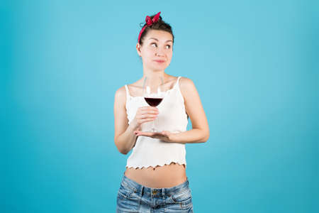 Young woman holds a glass of wine with two hands and looks away with a modest smile on a blue backgroundの写真素材