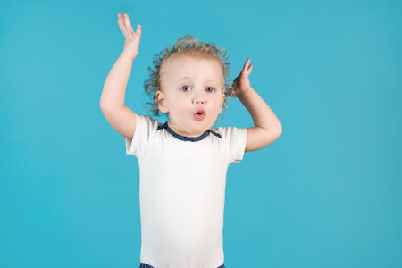 Funny curly baby girl of two years makes a surprised face and raises her hands up on a blue backgroundの写真素材