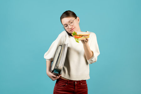 A girl with glasses with documents under her armpit is talking on the phone and is about to eat a sandwich. Looking at a sandwich with a smileの写真素材