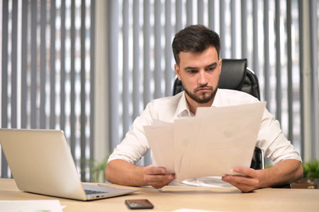 The young head of the company examines the reports printed on paper and is perplexed. Young boss is not happy with this seasons performanceの写真素材
