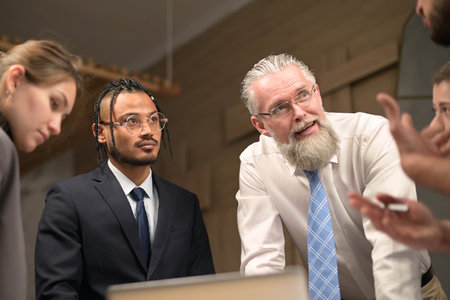 A mature solid gray-haired head of a firm discusses tasks with his subordinates. People of different nationalities and ages work together in a teamの写真素材