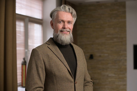 Stylish gray-haired man with a beard in a jacket looks confidently at the camera and smiling softly at homeの写真素材