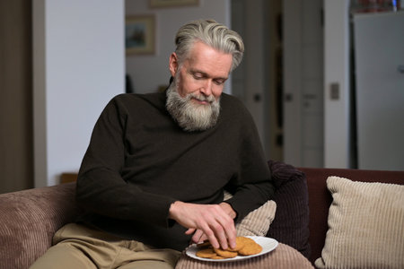 An adult gray-haired man with a beard sits at home on a sofa and eats a cookie, enjoyingの写真素材