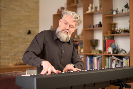 adult solid gray-haired man with a stylish beard plays the electronic piano at home in his spare timeの写真素材