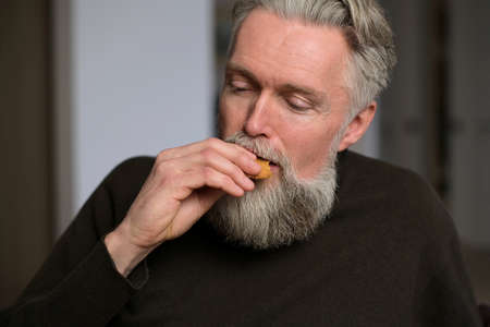 An adult gray-haired man with a beard sits at home and eats a cookie, enjoying
