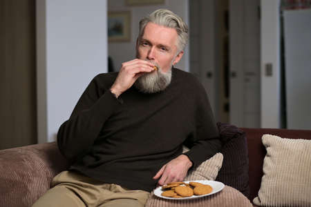 Adult gray-haired man sitting on the couch at home and eating cookiesの写真素材