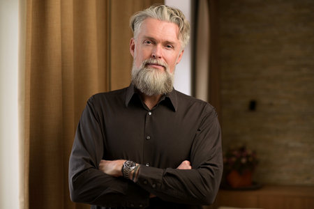 Stylish gray-haired man with a beard in a jacket looks confidently at the camera at homeの写真素材